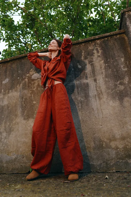 Cheerful woman outdoors in a loose red jumpsuit leaning against a weathered wall, sunlit moment, relaxed posture, natural tones, urban greenery in the background, lifestyle portrait