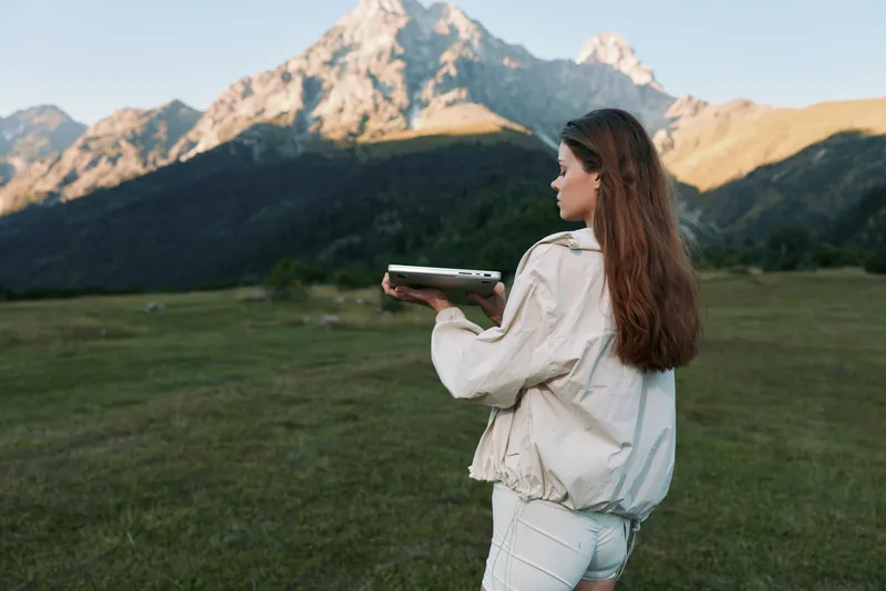 A woman stands in a meadow among distant mountains, holding a device in both hands. She looks toward the horizon with calm focus, embodying outdoor adventure.