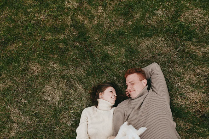 Happy couple lying on grass in cozy sweaters, smiling at each other, relaxed outdoor lifestyle moment with a small white dog nearby, warm intimate mood on a sunny day