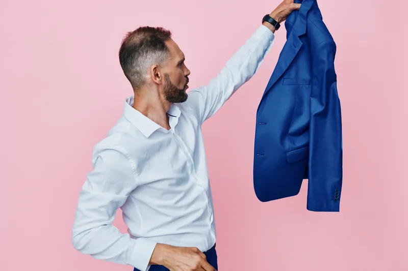A professional man lifts a vivid blue blazer against a soft pink background, capturing a moment of confident emotion, focus, and readiness in a polished business scene.