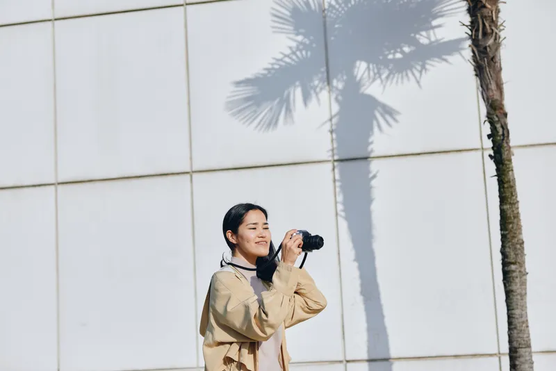 Photographer with camera capturing a moment outdoors, wearing a beige jacket beside a modern wall and tall palm trees, sunlight casting soft shadows for a candid, energetic outdoor filming scene