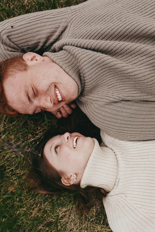 Cheerful couple outdoors, lying on grass, smiling with closed eyes, cozy sweaters, sunlit moment, relaxed posture, natural colors, gentle breeze, warm connection, serene countryside
