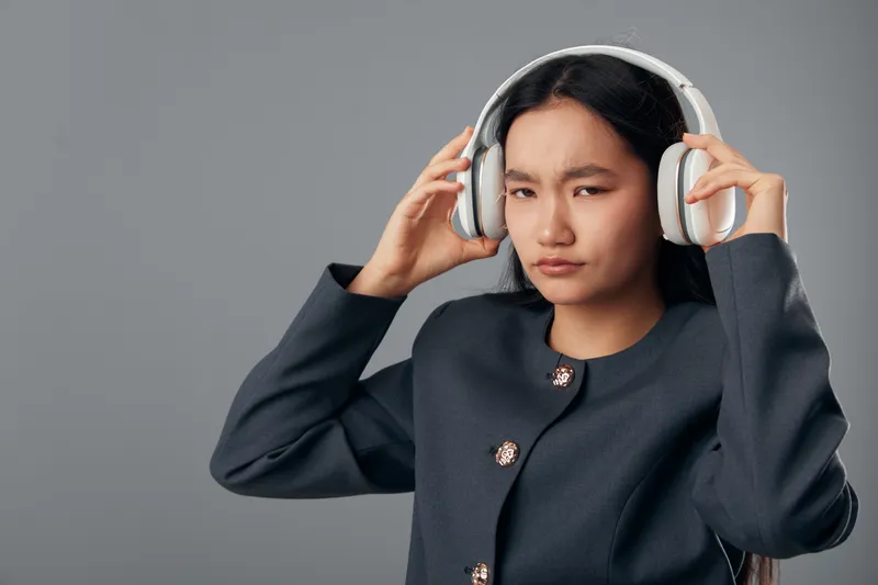 Young woman adjusting headphones with focused expression on gray background, wearing dark jacket, listening to music or audio with modern wireless headset technology.