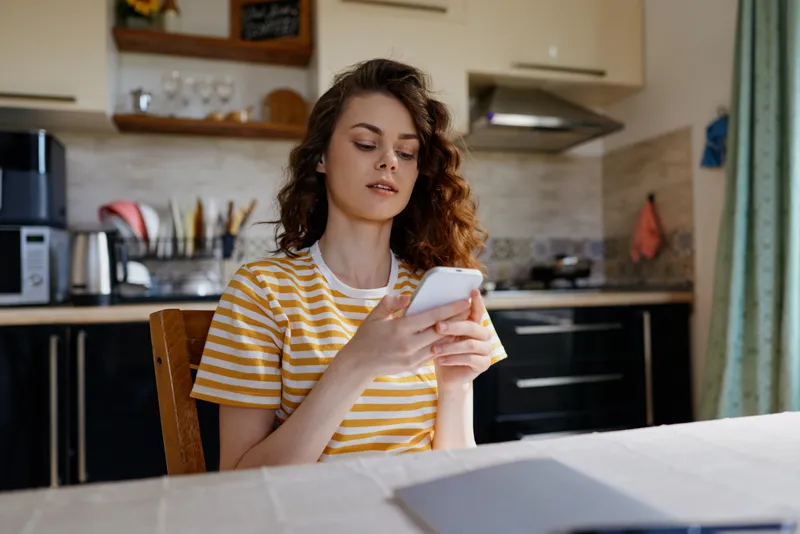 A young woman sits at a kitchen table, focused on her smartphone, wearing a striped tee, warm daylight in the home. A relaxed everyday moment highlighting digital routines.