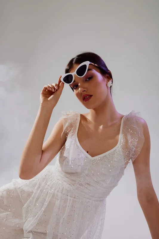 Young woman in a white elegant dress wearing stylish sunglasses poses confidently against a neutral background. Concept of beauty fashion and modern style.