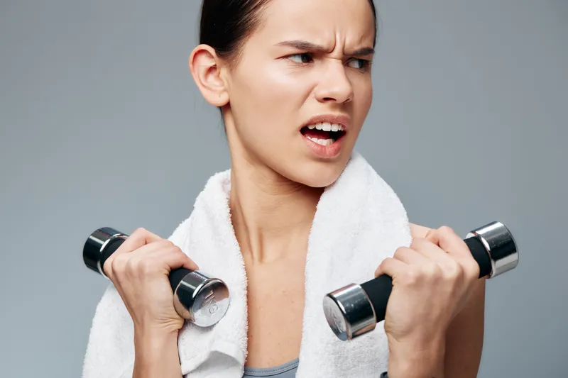 Athletic woman in gym holds dumbbells with a towel draped over her shoulders, intense expression as she powers through a strength workout, showcasing fitness and dedication.