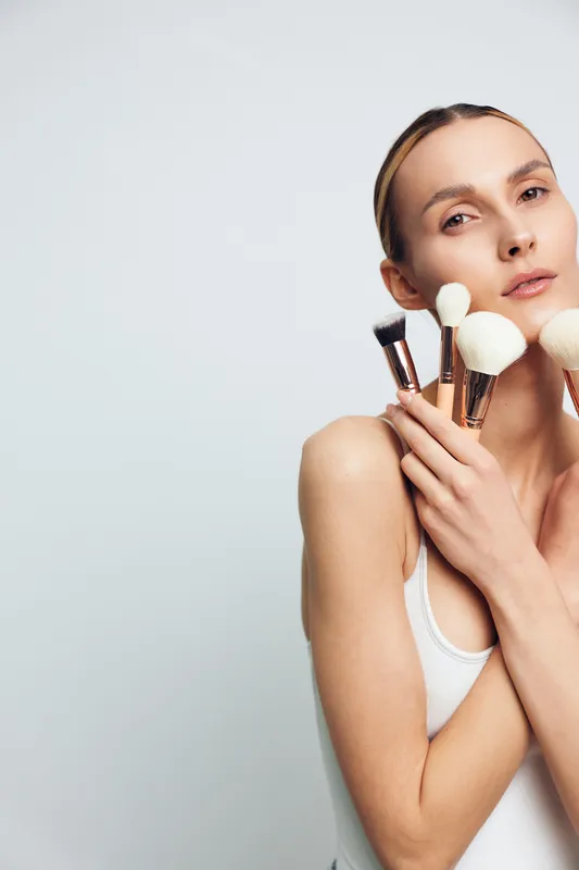 Young woman applying skincare cream to face with makeup brush, holding white sponge, calm beauty portrait, spa hygiene routine, clean background, softly copy space