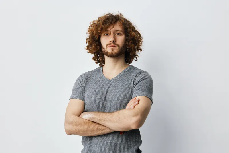 Confident young man with curly hair in a grey t shirt standing against a neutral background, conveying strength and self assurance