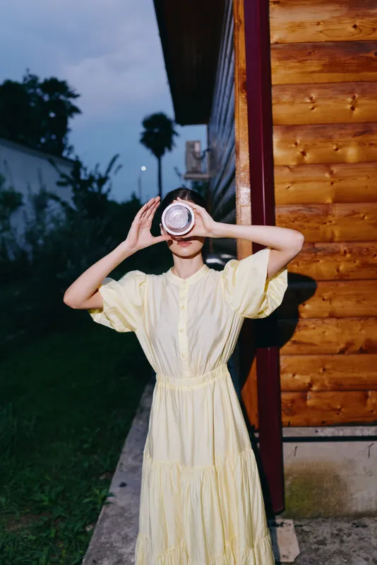 Woman in a pale yellow dress stands outdoors beside a wooden building, holding a camera to her eye and photographing the evening scene.