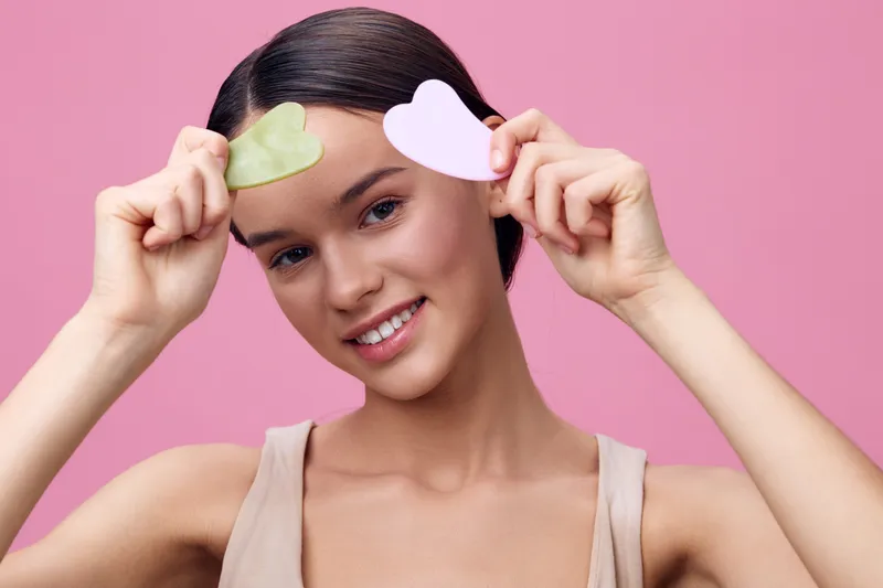 Young woman holding heart shaped skincare patches, smiling, against a soft pink background, embodying self care and beauty concepts emphasizing skincare and healthy routines
