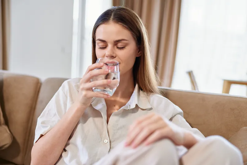 Woman sitting on sofa at home drinking a glass of water, relaxing with eyes closed in casual clothing, portrait of hydration, wellness and calm domestic morning routine and comfort.