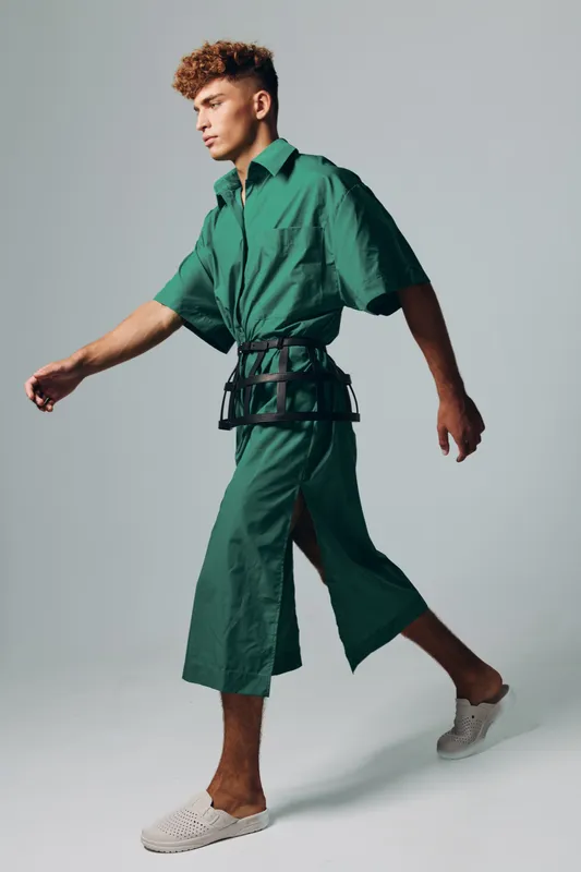 Young man walking in modern casual outfit with loose wide pants and a short sleeve shirt green color complemented by fashionable waist accessory. Stylish pose on light gray background in studio.