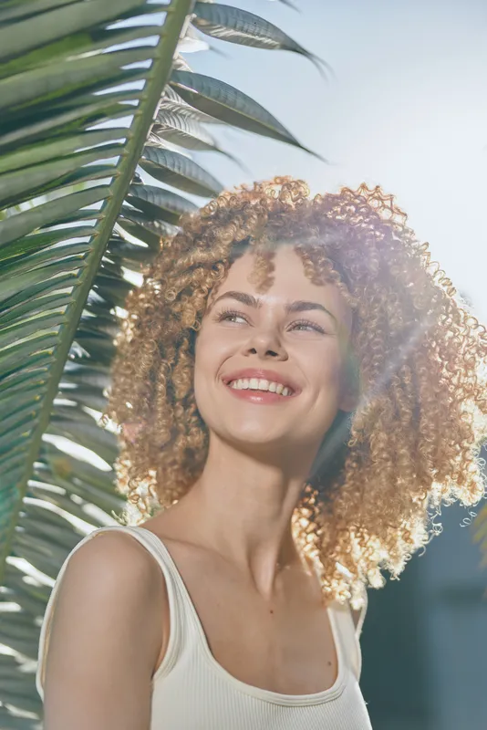 smiling woman with curly hair enjoys sunny tropical outdoors near large palm leaves, radiating happiness and natural beauty in summer light.