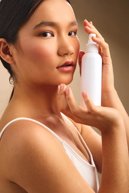 asian woman holds a white cosmetic bottle near her face in a soft studio setting, highlighting skincare and beauty routine with a calm, confident expression and warm, appealing light.