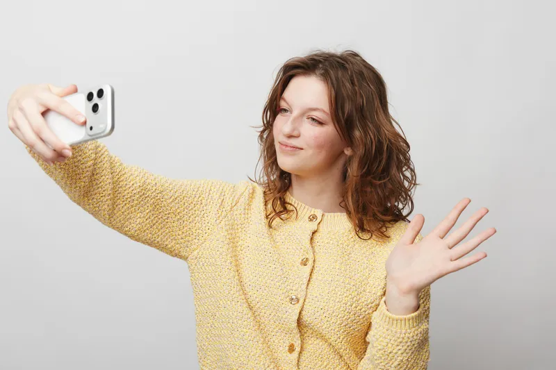 Young woman with curly hair wearing yellow sweater takes selfie with smartphone and waves hand. Bright studio background. Concept of social media, communication, and casual lifestyle.
