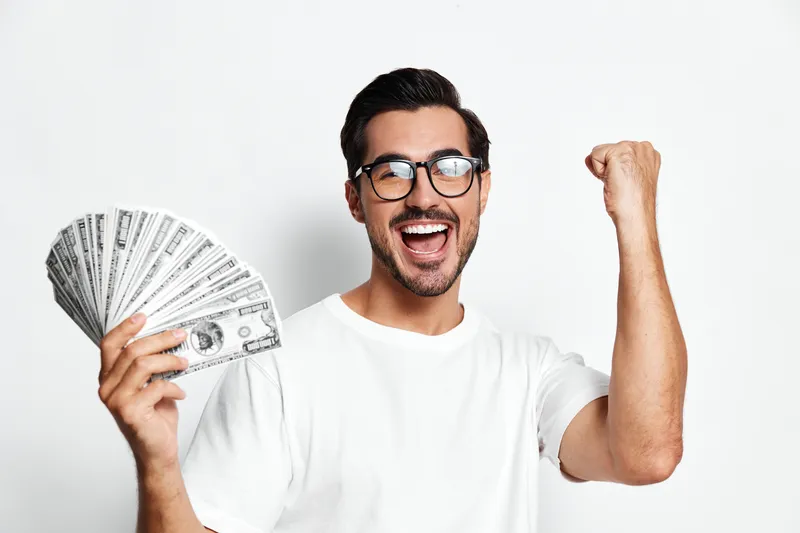 Happy cheerful man in glasses celebrating success, holding fan of money while standing against plain white background. Financial success concept.