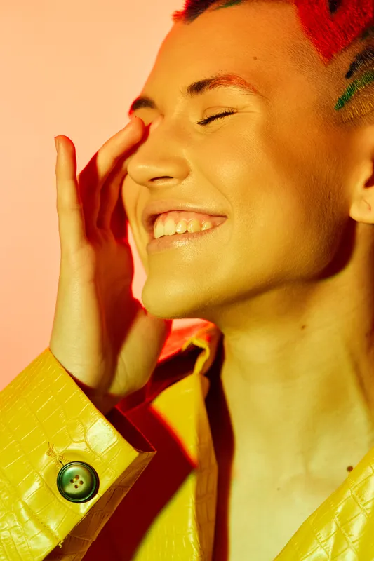 Smiling woman with colorful hair and yellow jacket expresses joy and positivity against warm orange background, radiating vibrant energy and happiness.