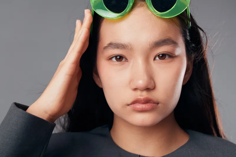 Young woman with long dark hair and neutral expression wearing green sunglasses on her head against gray background. Portrait of stylish female fashion model with natural makeup looking at camera.