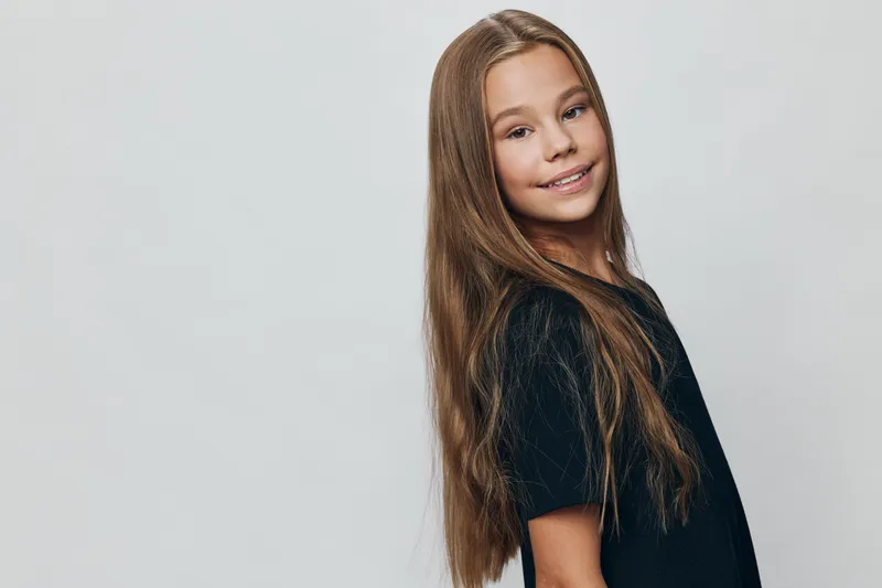 Girl with long brown hair smiling and posing in black t shirt with natural expression against grey background in studio for portrait and youth fashion concept.