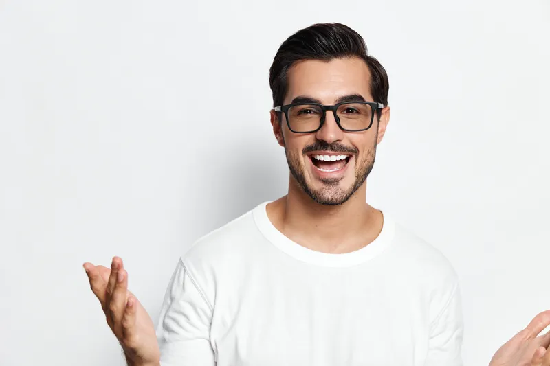 Happy smiling young man with glasses joyfully engaging with the camera, wearing a casual white t-shirt, isolated on a solid light background. Lifestyle concept.
