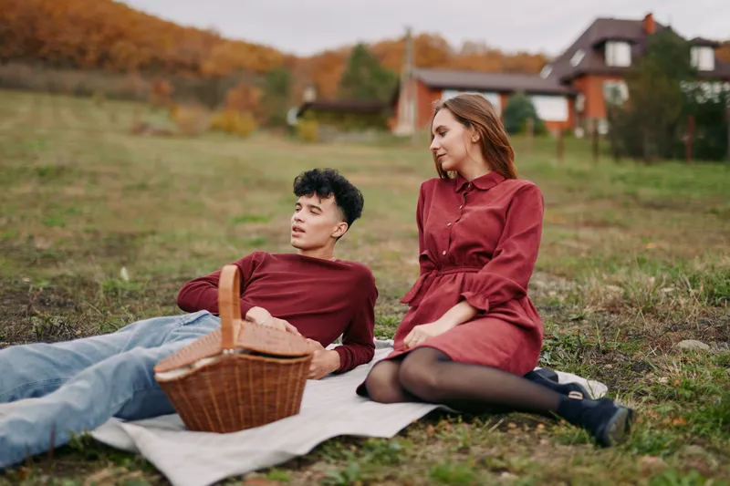 A genuine outdoor picnic scene showing an authentic young couple relaxing on a blanket in a rural meadow, natural gestures and candid expressions conveying honest connection.