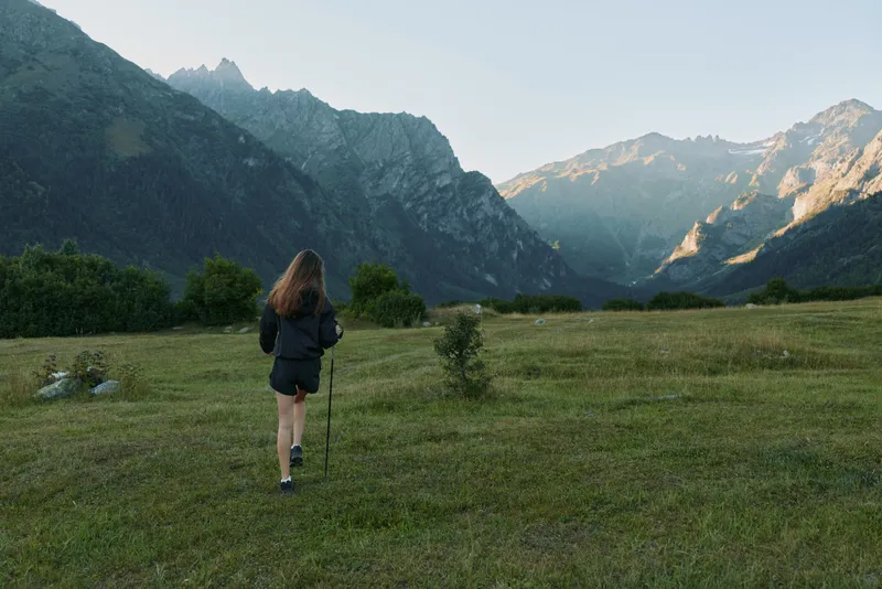 A hiker walks across a sunlit meadow toward dramatic mountain peaks, backpack resting on shoulders, conveying exploration, solitude, and a tranquil outdoor adventure.