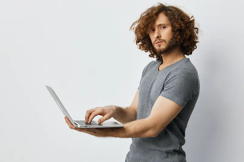 Young man with curly hair working on a laptop, wearing a gray t shirt, displaying focused expression against a light background