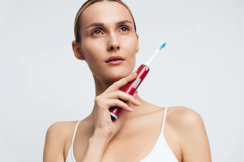A thoughtful woman in a white tank top holds a red electric toothbrush near her face, looking upwards against a plain light background. Concept for dental care and personal hygiene.