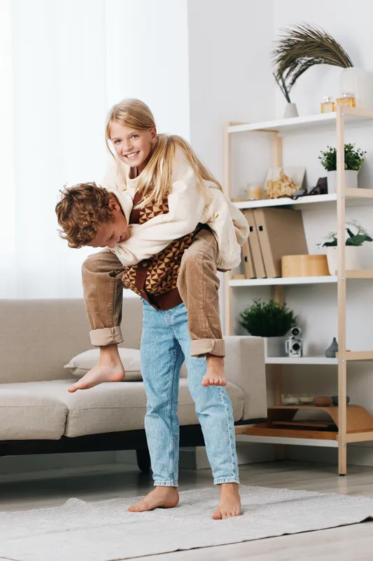Authentic moment of joyful play in a bright home, showcasing a school themed scene and lifestyle balance as siblings share a piggyback ride on a soft living room rug.