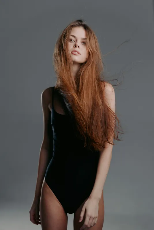 Young woman with long hair wearing black bodysuit posing indoors with neutral expression against grey background
