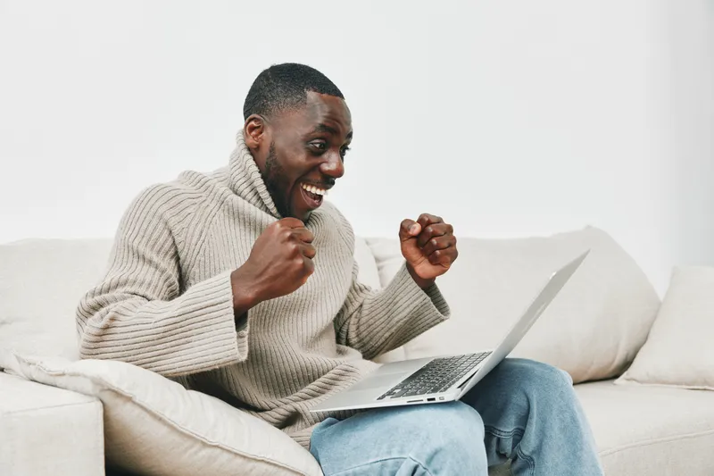 Happy African man celebrating success while using a laptop at home, wearing a stylish sweater in a cozy, light colored living room setting