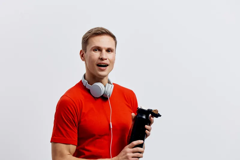 smiling young caucasian man wearing red t-shirt holding shaker bottle headphones around neck studio portrait fitness workout concept