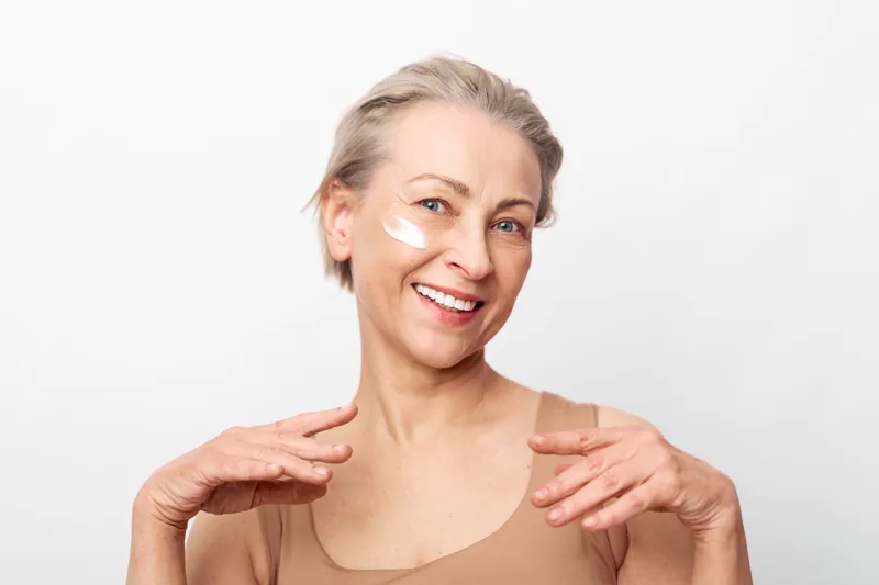 Smiling middle aged woman with short hair applies white face cream on cheek. Studio portrait on white background. Concept of skincare, anti aging, self care, beauty routine and wellness.