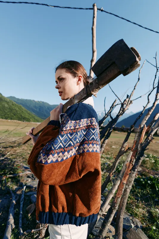 Woman with axe and patterned sweater in countryside mountains, standing by wooden fence in a rural portrait, outdoors scene with sunlight, meadow and rustic homestead vibe.