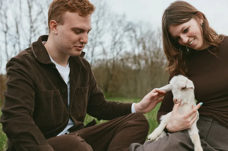 Young couple in casual clothes gently holds and pets a small white baby goat sitting on the woman's lap in a natural outdoor setting with bare trees in early spring.