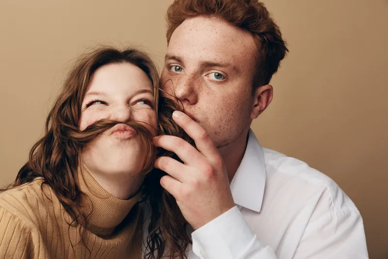 Young couple with red hair playfully posing against beige background. Man holds woman’s hair as mustache. Concept of fun, love, friendship, and casual lifestyle together.