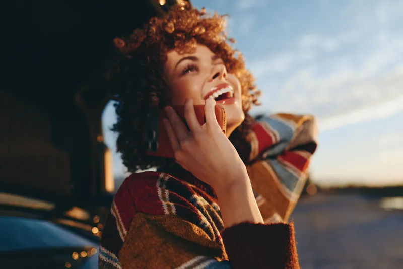 Woman lifestyle, phone smile, curly hair, outdoors sunlit sunset, warm communication, relaxed adult woman holding device, patterned jacket, cheerful expression, natural background