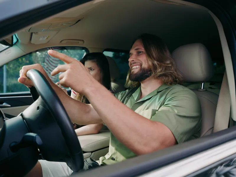 Joyful man driving a car, smiling with one hand on the steering wheel, a passenger beside him, bright interior, sense of momentum, casual travel vibe, daytime scene.
