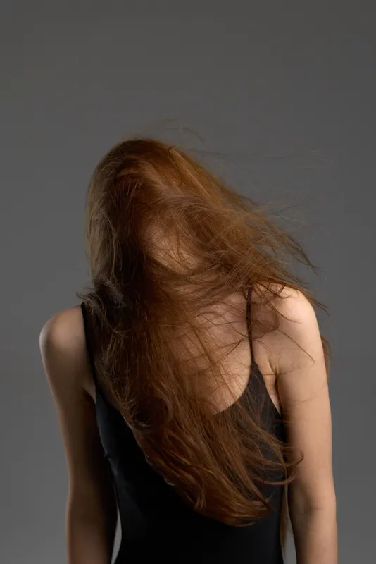 Woman with long red hair blowing over face wearing black tank top against gray background