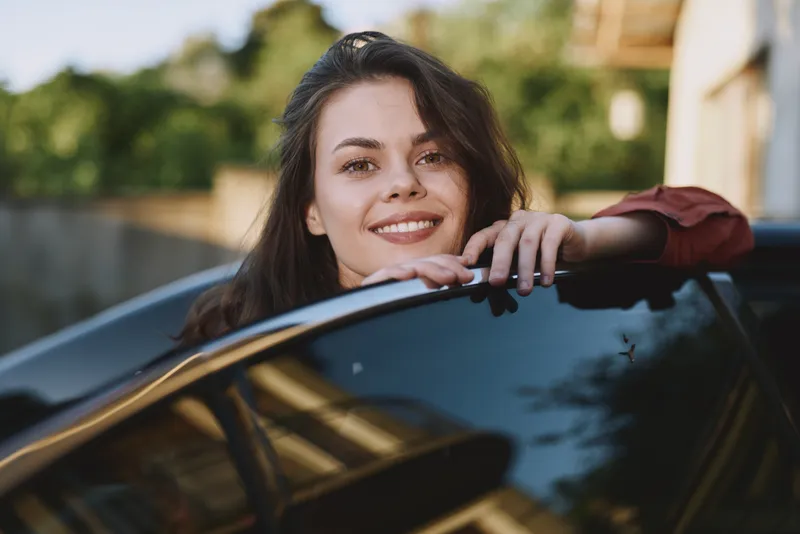 A young woman leans on a car window, smiling warmly in a sunlit street scene, conveying casual confidence and everyday happiness