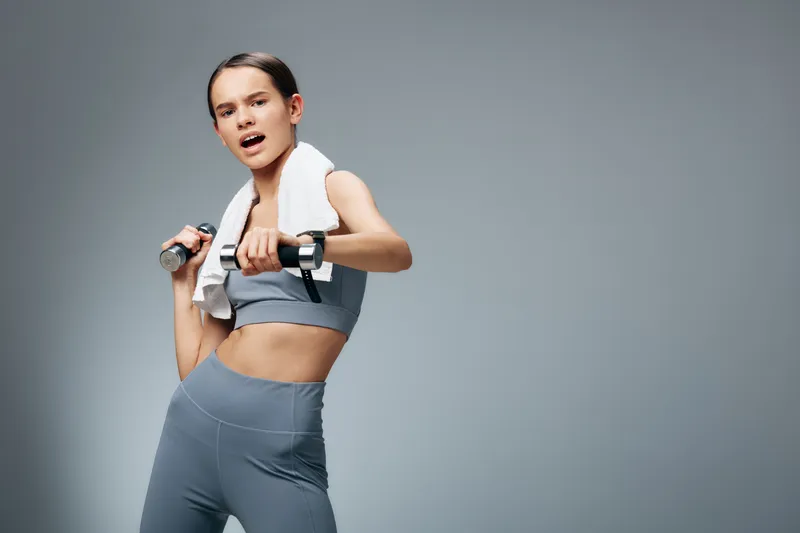 Energetic woman in athleticwear lifts dumbbells with a towel draped over shoulders, mid workout in a neutral studio, showcasing strength, focus and a healthy lifestyle.