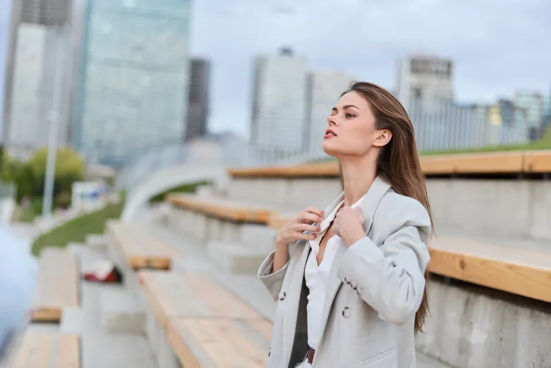 Confident woman in a business suit adjusting her collar outdoors with a modern city backdrop, embodying professionalism and elegance in a dynamic urban setting.