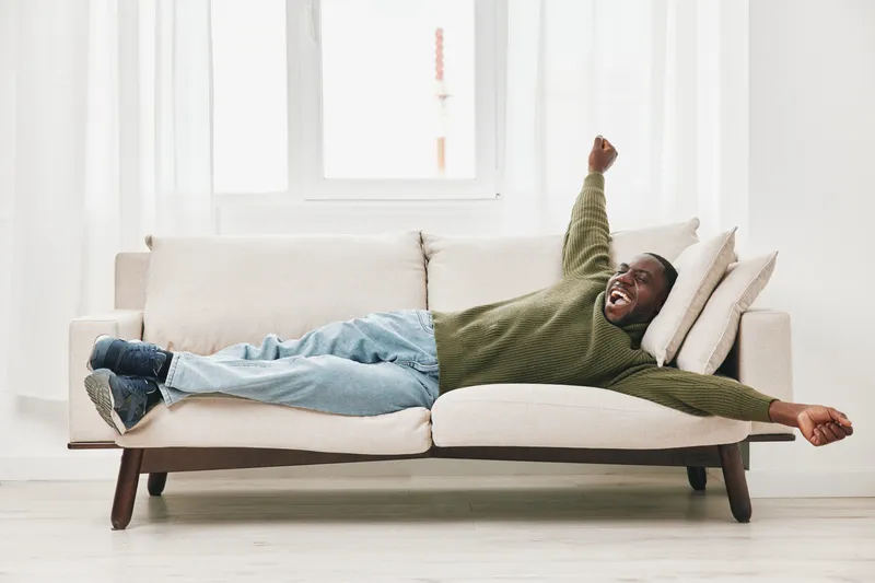 A joyful Black man relaxing on a couch, wearing a green sweater and blue jeans, embodying comfort and happiness in a bright, airy living space