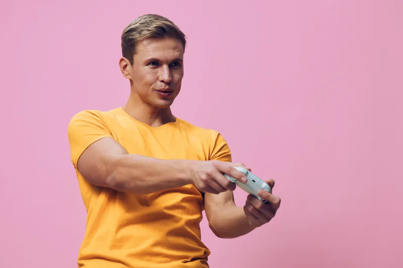young man in yellow t-shirt playing videogame holding joystick isolated on pink background focused digital entertainment concept