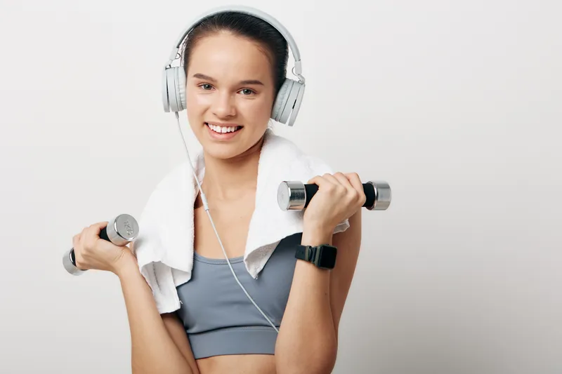Woman in athletic wear with a towel around her neck, headphones on, and dumbbells in hand, mid workout pose showcasing a fitness routine, strength training and focus in a bright studio.