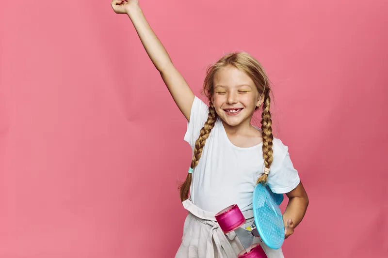 Happy young girl with braided hair holding a pink skateboard raises her arm with closed eyes smiling. Studio shot on pink background. Concept for childhood and joy.