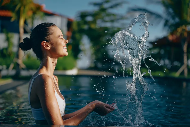 Young woman in white swimsuit enjoys refreshing water splash at outdoor pool with tropical background sunny day smiling relaxed lifestyle concept.