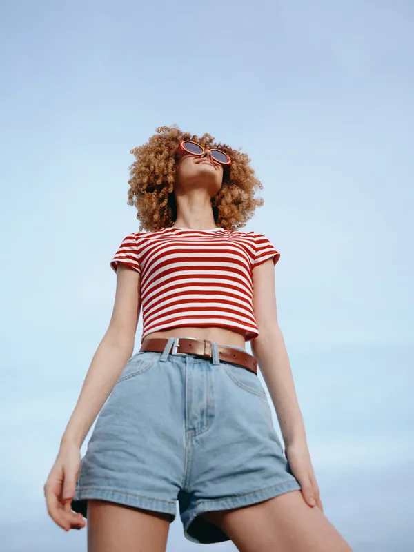 A stylish young woman stands outdoors under a clear blue sky, wearing a red and white striped crop top, high waisted denim shorts, and sunglasses, posing confidently with wind in her hair.