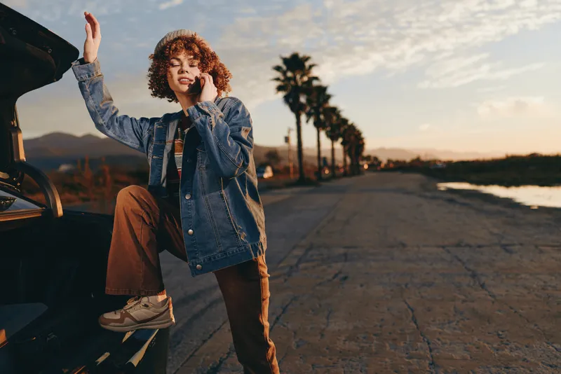 Woman lifestyle outdoors sunset denim jacket, reaching toward the sky beside a parked car on a road, curly hair and casual pose in warm golden light, fashion portrait traveling freedom