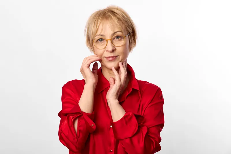 Woman with glasses smiling portrait, mature expression as hands touch face, red shirt on a white studio background, closeup conveys calm beauty, wellbeing, and self care